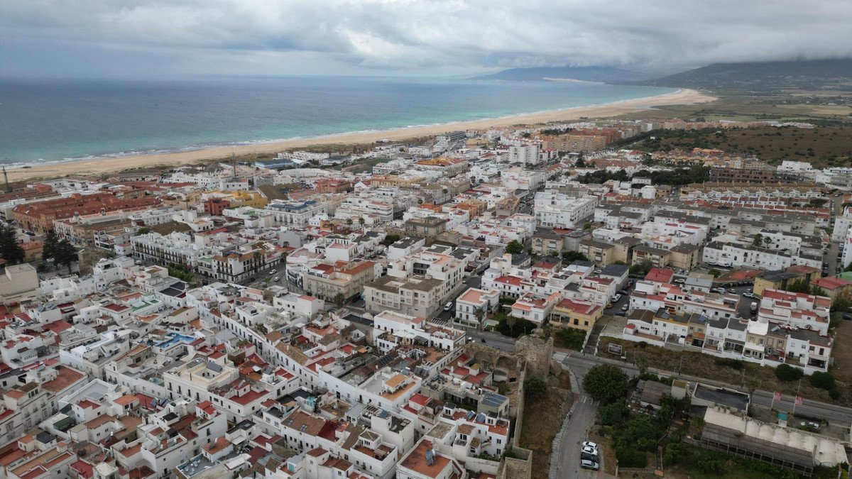 Aerial view of a coastal town with whitewashed buildings, a sandy beach, and the sea under a cloudy sky.