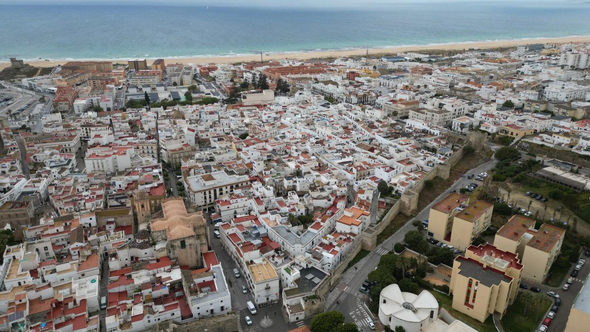 Aerial view of a coastal town with white buildings, red rooftops, and a beach meeting the blue sea under an overcast sky.