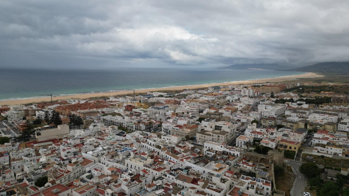 Aerial view of a coastal town with white buildings, a long sandy beach, and the sea under a cloudy sky.