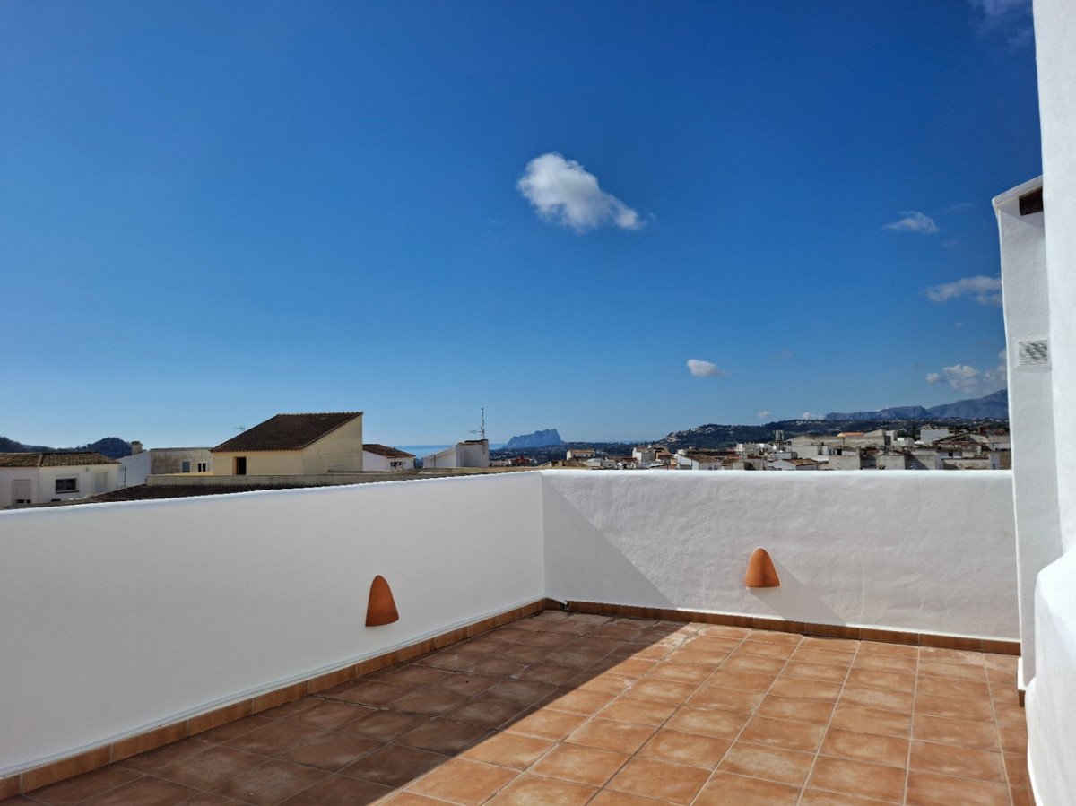 A sunny rooftop terrace with tiled flooring and white walls, overlooking a town and distant mountains.