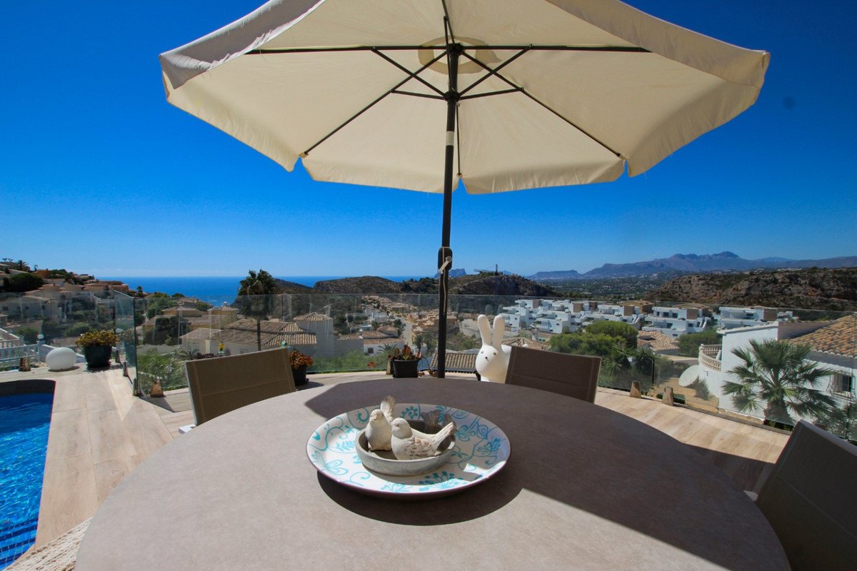 Terrace with dining table, chairs, umbrella, and a pool, overlooking a town and the sea.