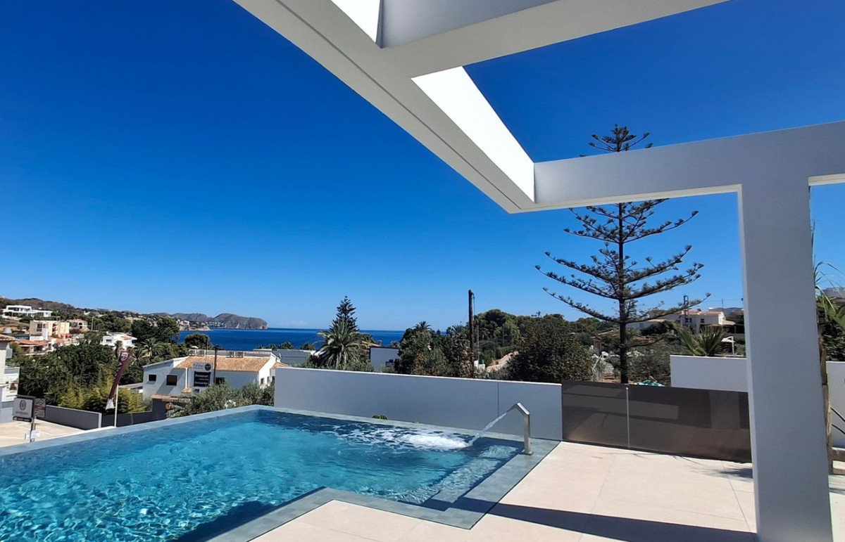 Modern infinity pool with a cascading water feature on a terrace, overlooking the sea and coastal town on a clear day.
