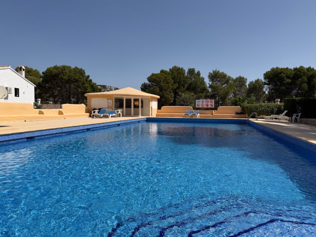 A large blue swimming pool with surrounding lounge chairs and a small building in the background, under a clear blue sky.