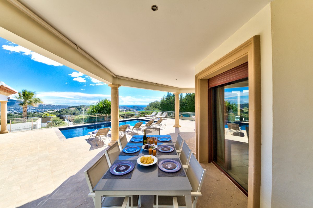 Covered patio with dining table set for a meal, overlooking a swimming pool and the sea with a panoramic sea view.