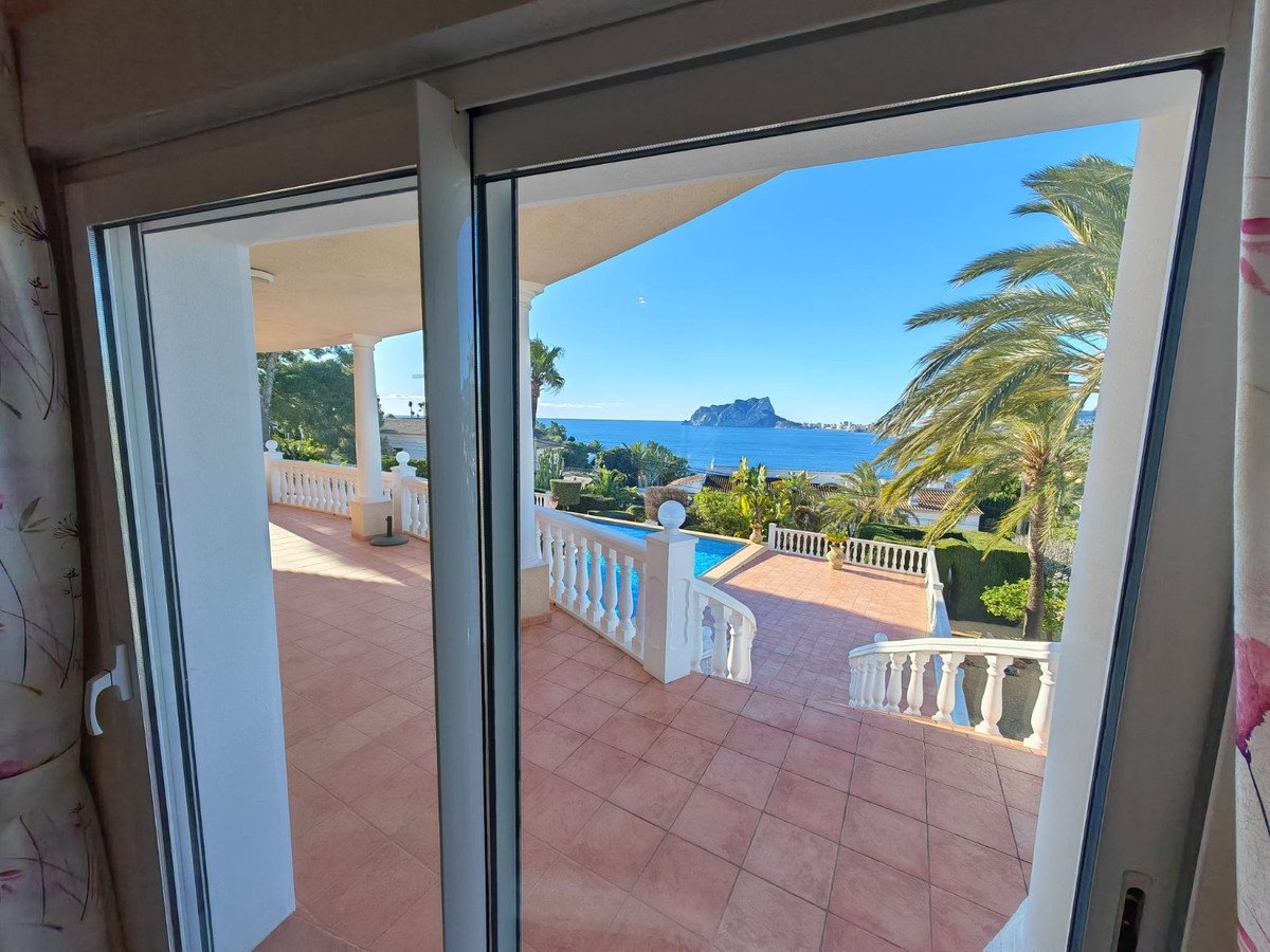 View from sliding doors onto a tiled terrace with a pool, palm trees, and a distant sea view with a mountain.