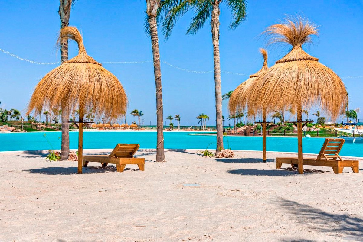 Two lounge chairs under thatched umbrellas on a sandy beach beside a large, bright blue swimming pool with palm trees and clear sky.