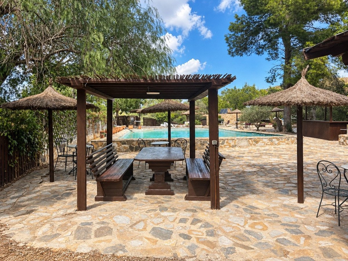 Outdoor wooden dining area with benches and tables beside a swimming pool under a pergola and straw umbrellas.