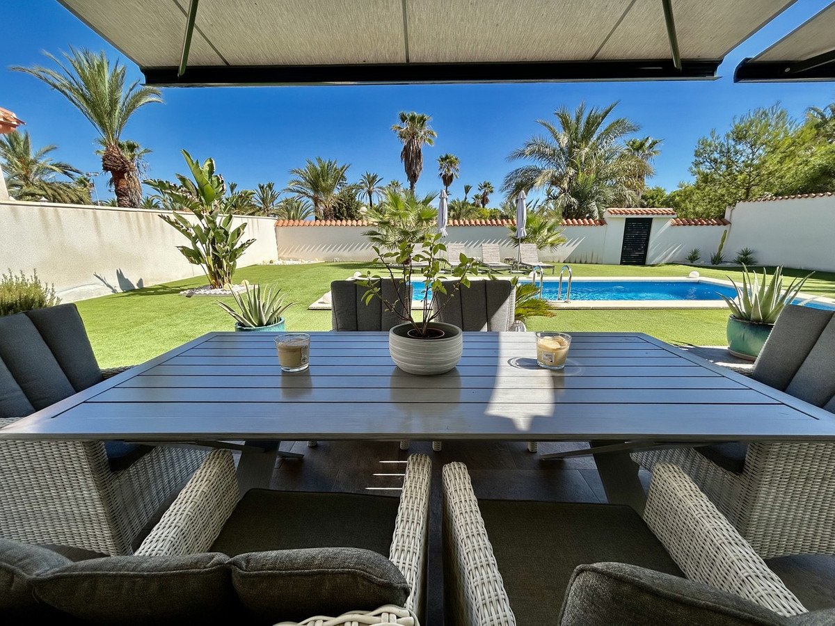 Patio dining area with outdoor furniture overlooking a sparkling blue swimming pool and lush green garden with palm trees.