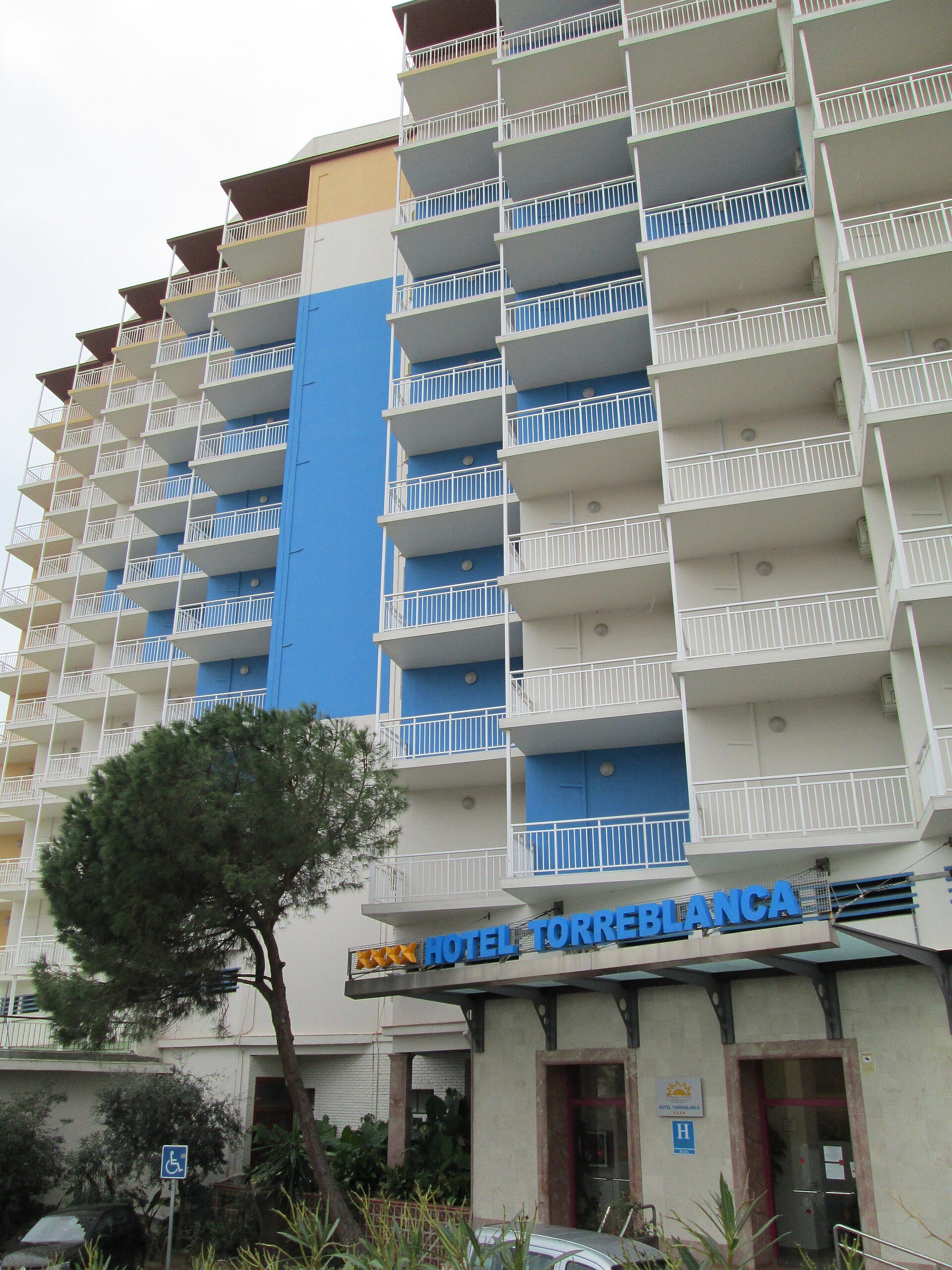 Multi-story hotel with balconies, blue and white exterior, pool, and sea view.