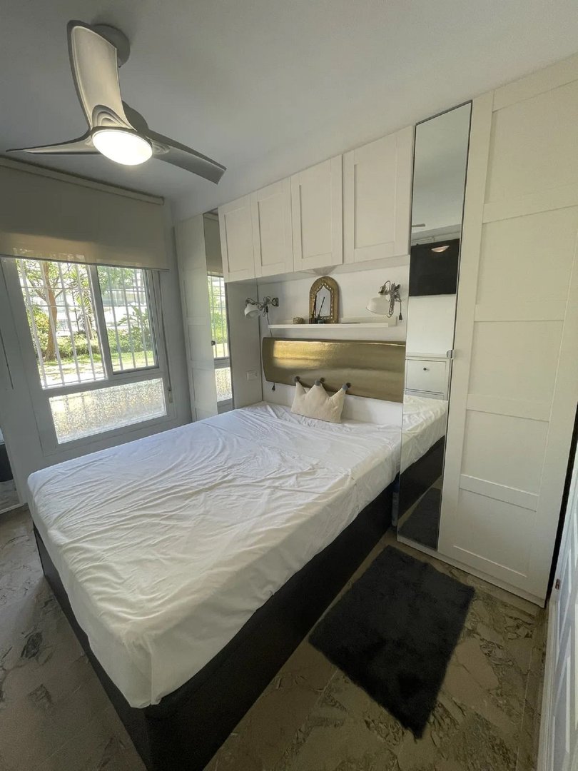A bedroom with a bed, built-in white wardrobes with mirrored doors, and a window overlooking trees. A ceiling fan is visible.