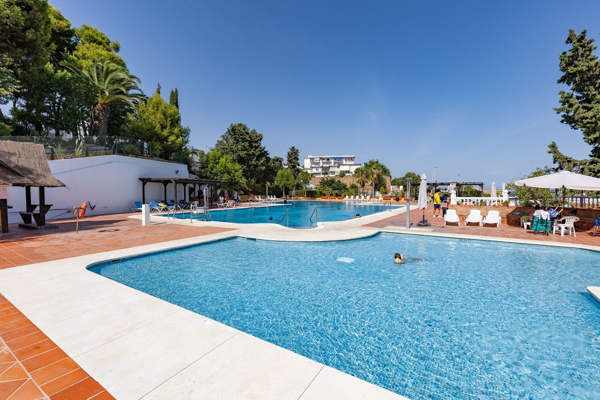 Large outdoor swimming pool with loungers and umbrellas under a clear blue sky, surrounded by trees and buildings.