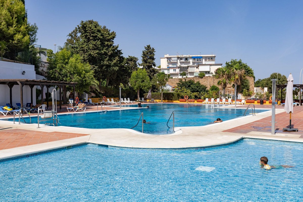 A large outdoor swimming pool complex with sunbeds, umbrellas, and lush trees surrounding it, under a clear blue sky.