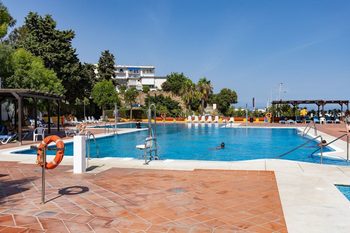 Large outdoor swimming pool with sun loungers, umbrellas, and people swimming under a clear blue sky.