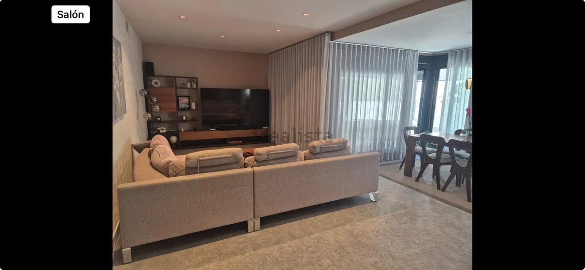 A modern living room with a large sectional sofa, TV, and a dining area with a wooden table and chairs visible through sheer curtains.
