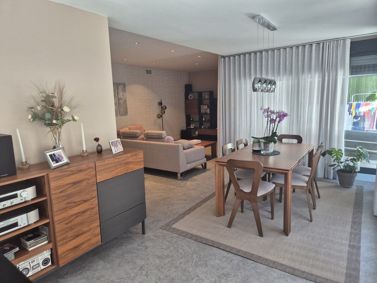 A modern dining room with a wooden table and chairs, adjacent to a living area with a sofa. Curtains cover the windows.