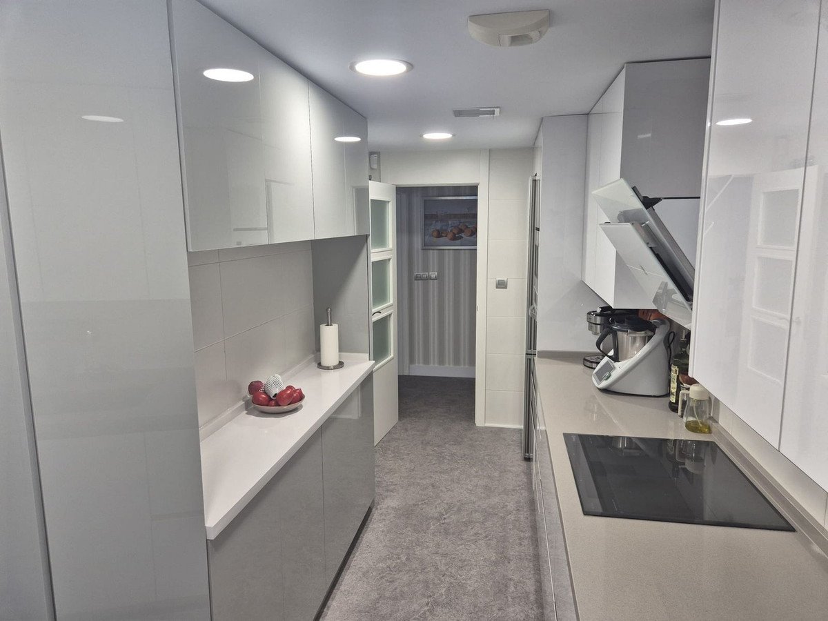 A modern, narrow kitchen with white glossy cabinets, gray countertops, and a built-in stovetop and hood.