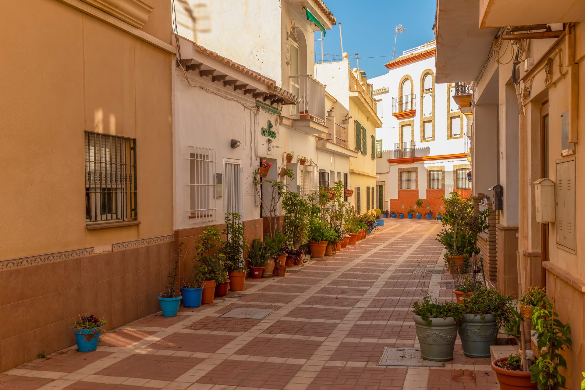 Narrow alley with potted plants, white and beige buildings, clear blue sky.