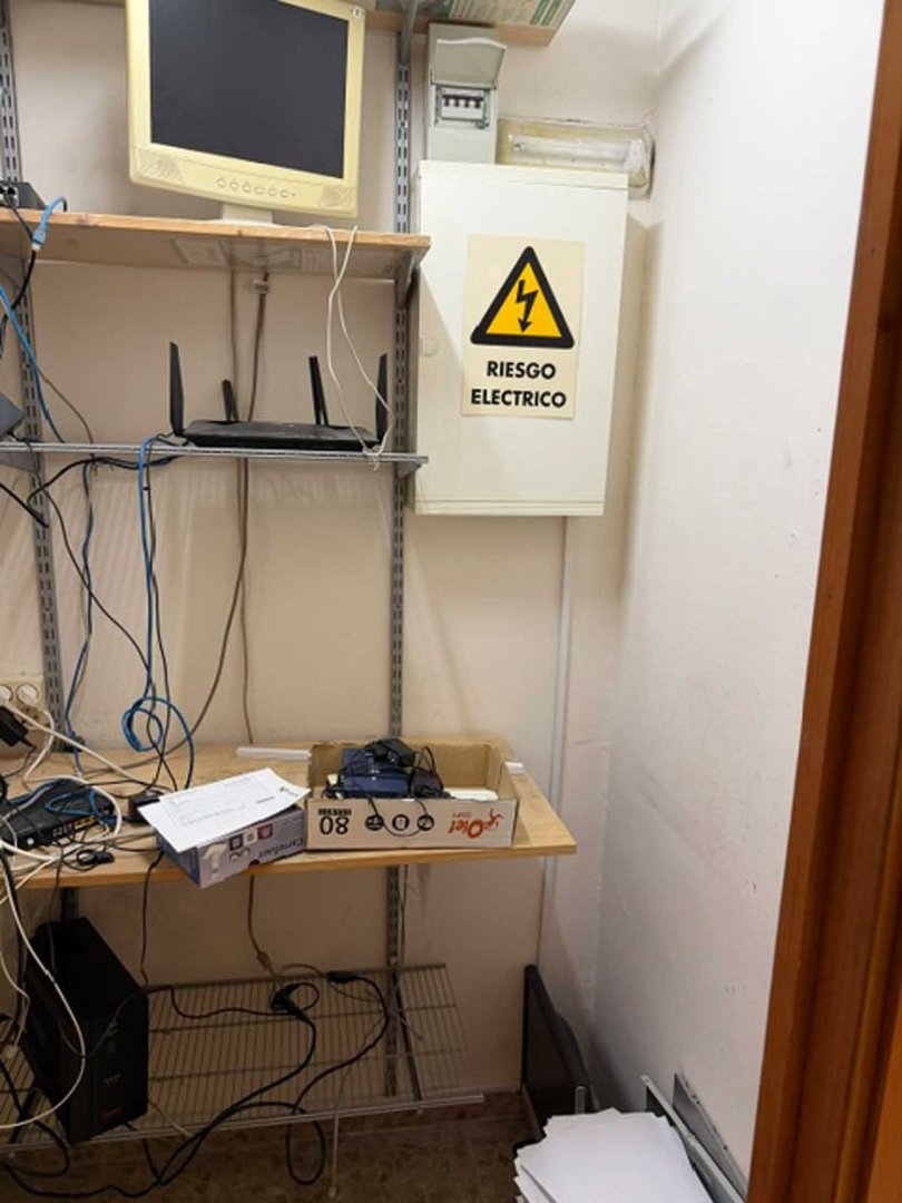A small room with shelves holding networking equipment, computers, and a monitor. A high voltage warning sign is visible.