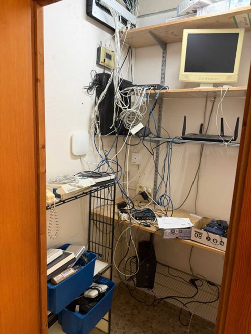 A messy server room with computer equipment, routers, and tangled wires on shelves.