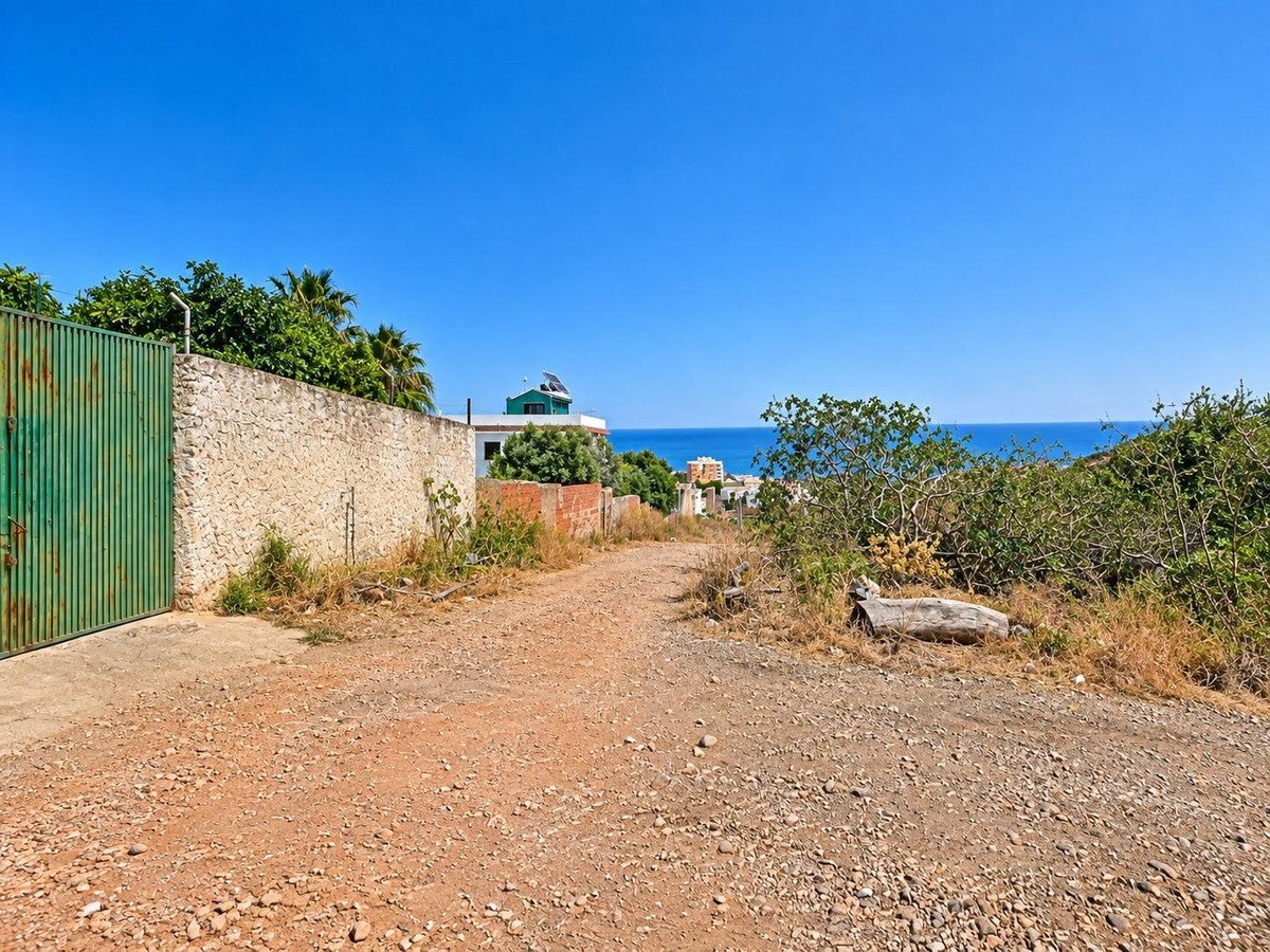 A dirt pathway leading towards the sea, with buildings and lush greenery visible in the distance under a clear blue sky.