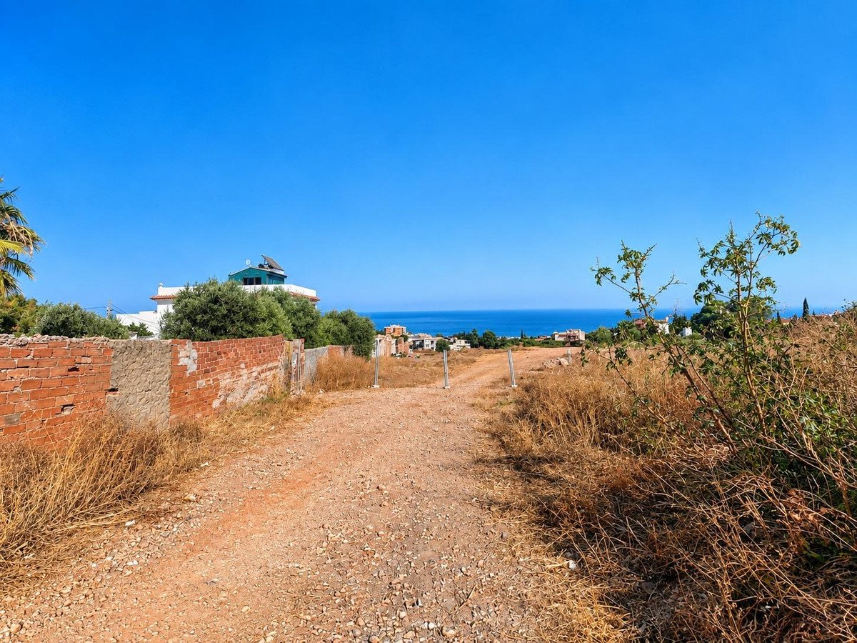 A dirt road leads towards a distant sea view, with houses and trees visible along the coastline under a clear blue sky.