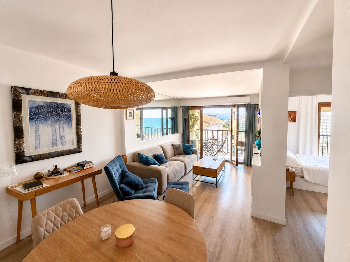 Living room with a beige sofa, blue accent chair, and a dining table. Sliding doors open to a balcony with a sea view.