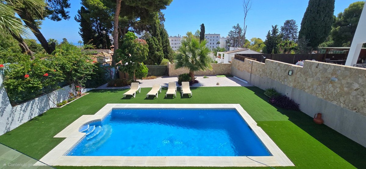 Outdoor swimming pool with sun loungers and artificial grass garden, featuring palm trees and surrounding greenery.