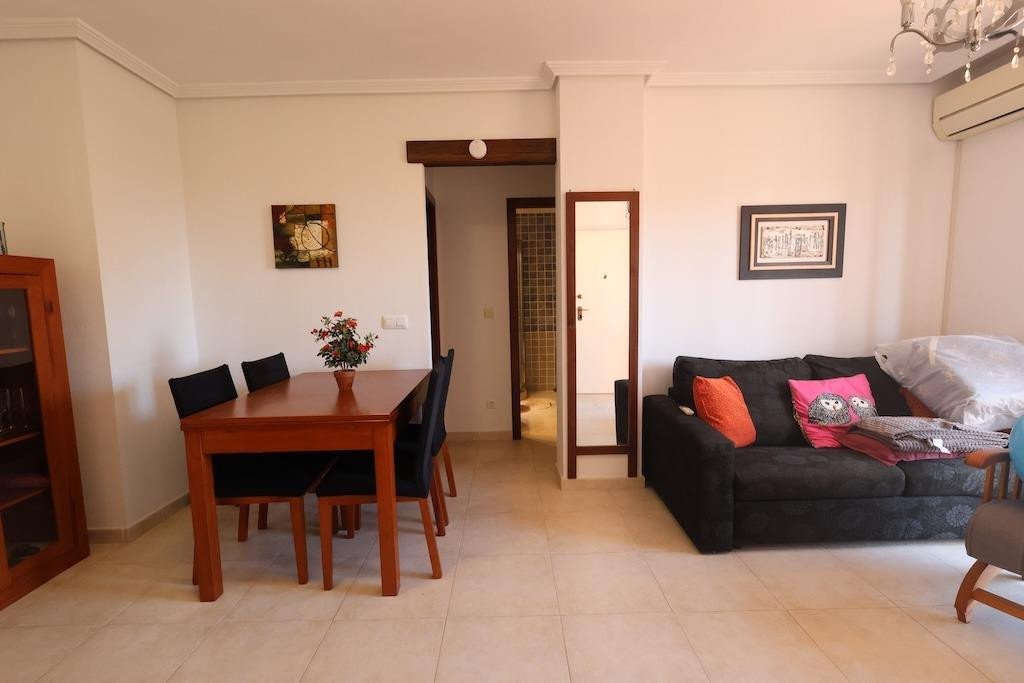 A dining area with a wooden table and chairs next to a living area with a dark sofa, orange and pink pillows, and a mirror.