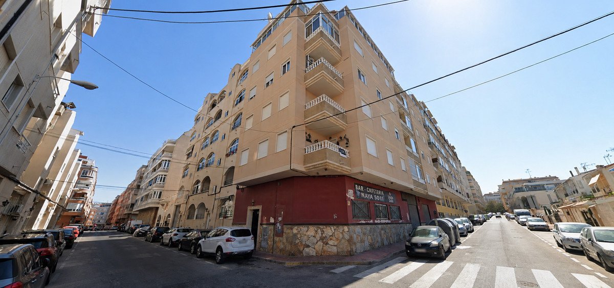 Exterior view of a tan apartment building with multiple balconies, situated on a street with parked cars and a bar on the ground floor.