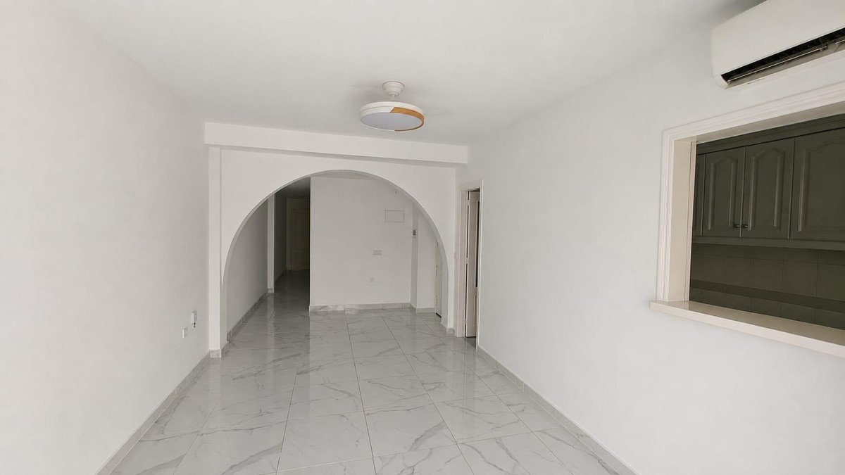 A bright living area with white walls, marble flooring, an arched doorway, and a view into a kitchenette with dark green cabinets.