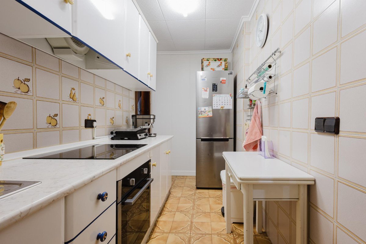 A modern kitchen with white cabinets, induction hob, oven, and a stainless steel refrigerator. Fruit-themed tiles are on the backsplash.