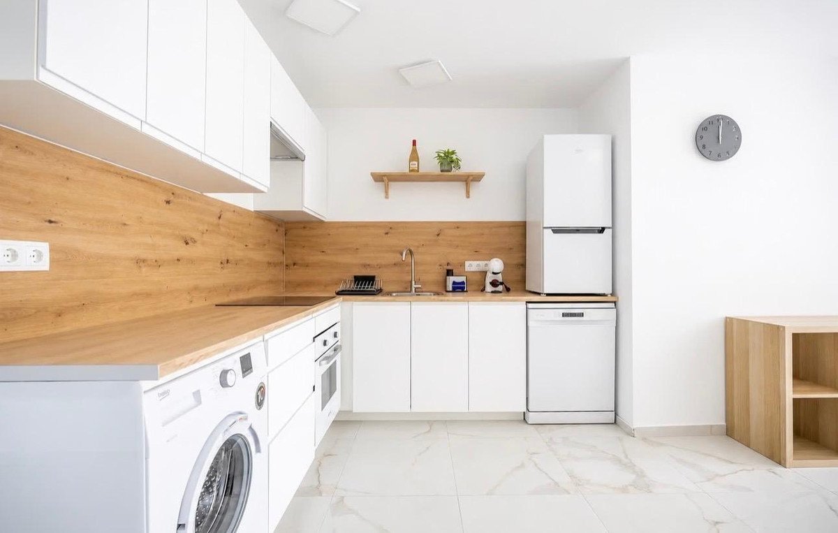 A modern white kitchen featuring wooden countertops and backsplash, stainless steel appliances, and white cabinetry. Includes a dishwasher, refrigerator, and washing machine.