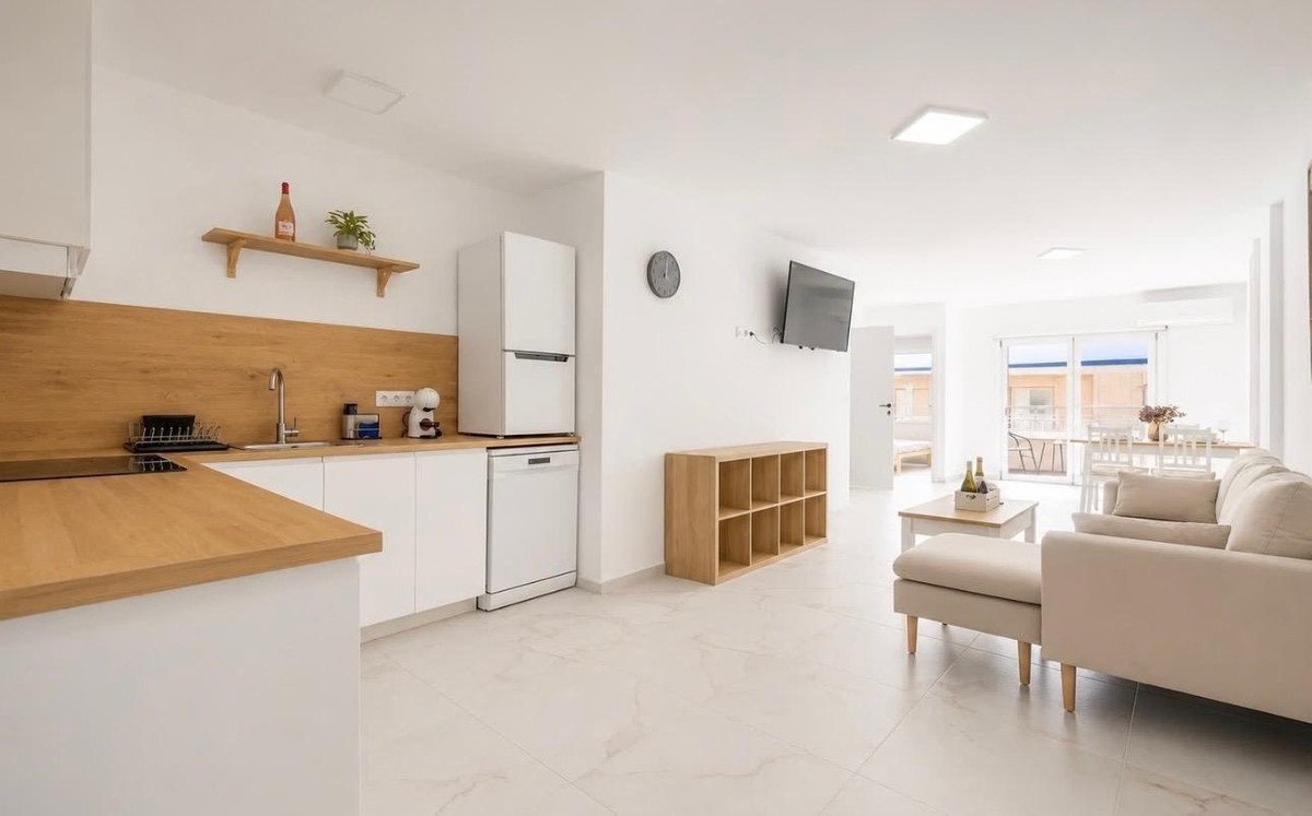 A modern open-plan living area with a kitchen, sofa, dining table, and large windows offering a view of the outside.