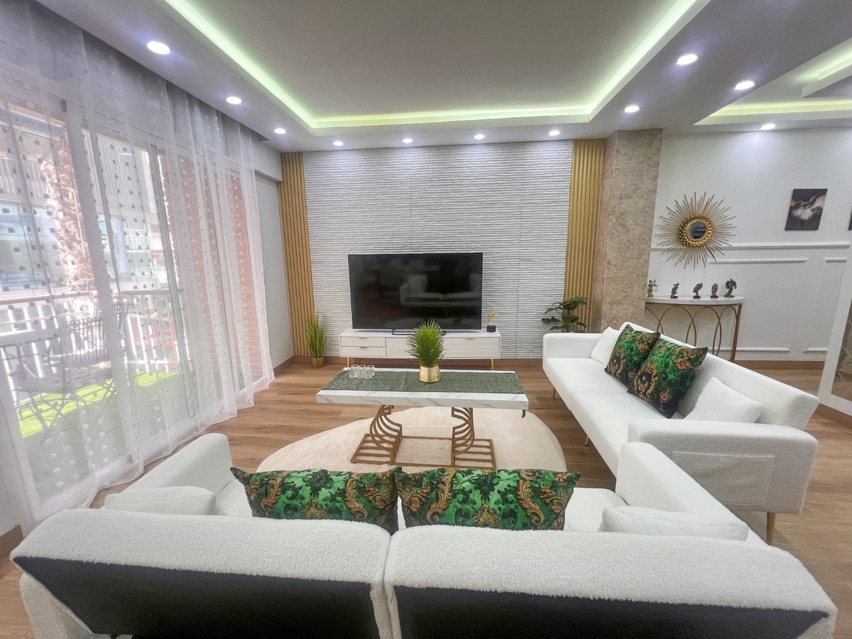 A modern white living room with comfortable sofas, a coffee table, a television, and a large window overlooking a balcony with greenery.