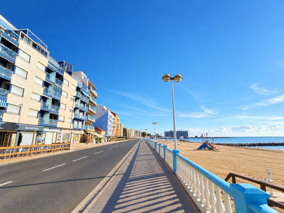 Apartment buildings line the street along a sandy beach with a blue railing and the sea under a clear blue sky.