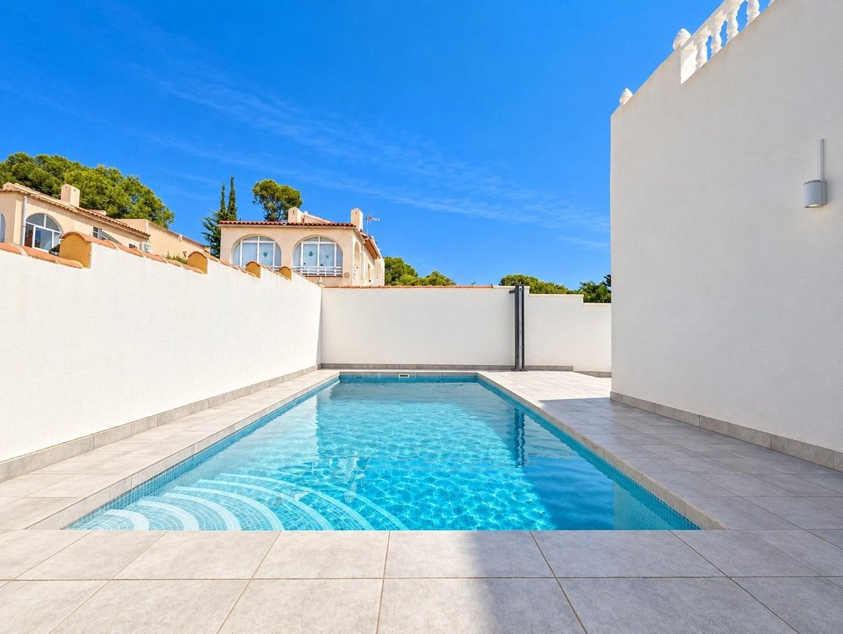A clean, rectangular swimming pool with blue water and steps, surrounded by a tiled patio and white walls under a clear blue sky.