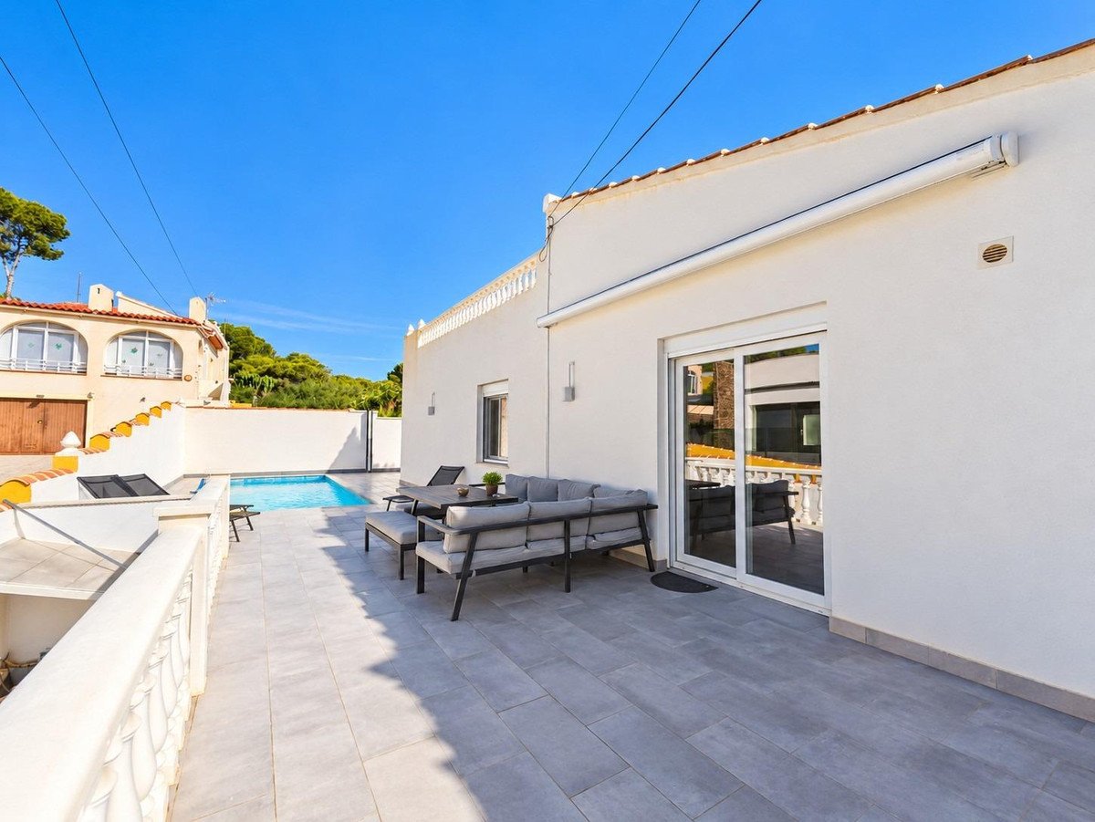 Modern white house with a tiled patio, outdoor seating, and a private swimming pool on a sunny day.