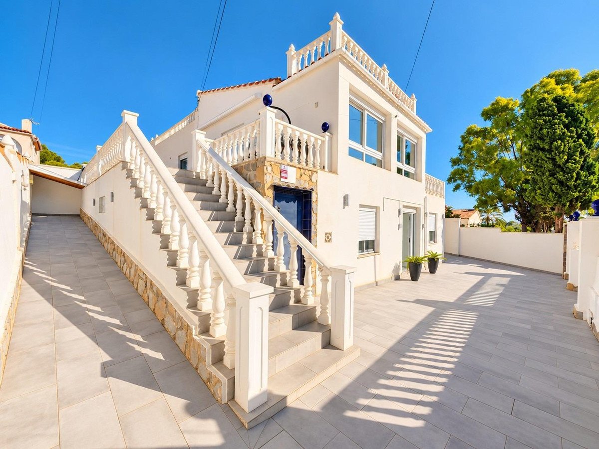 Exterior of a white villa with a grand staircase, terrace, and driveway on a sunny day with blue sky.