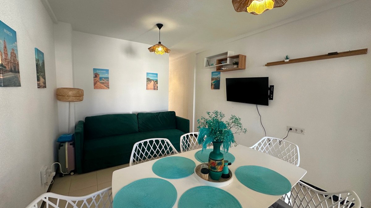 A modern living and dining area featuring a green sofa, white chairs, a dining table with turquoise placemats, and wall art on the walls.