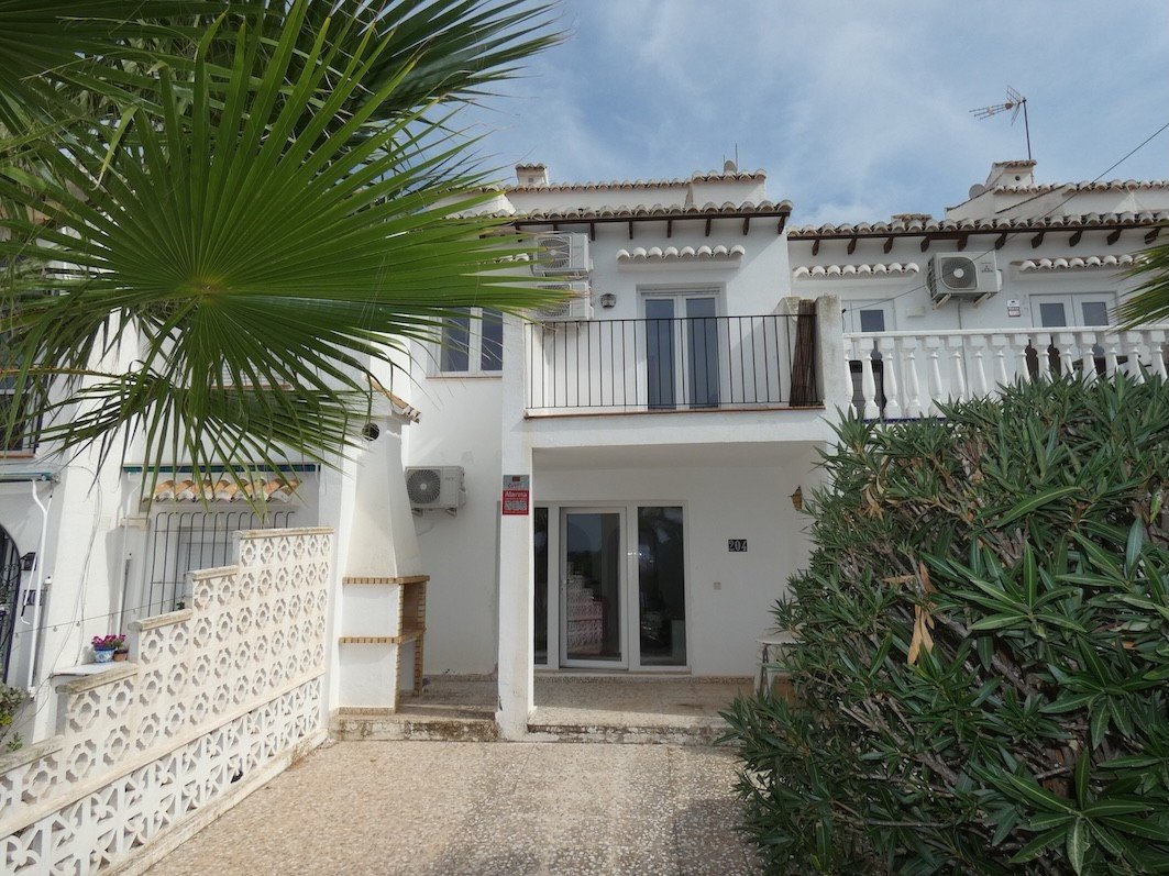 White townhouse with balconies, air conditioning units, and a patio area, partially obscured by palm leaves and greenery.