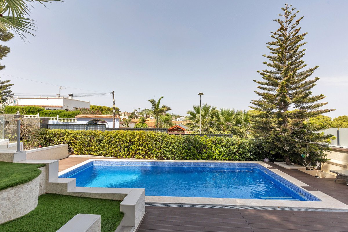 A bright blue swimming pool with tiled edges, surrounded by a wooden deck, green lawn, and lush hedges. Outdoor landscaping with a large pine tree.