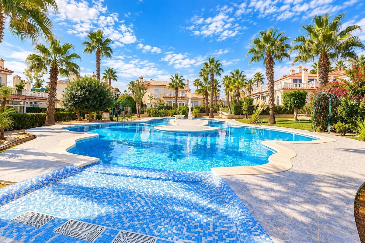 Spacious communal pool with blue tiles, surrounded by palm trees and residential buildings under a sunny sky.