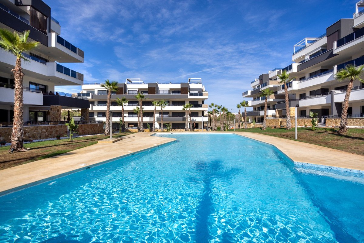 Modern apartment buildings with balconies surrounding a large, blue swimming pool and palm trees on a sunny day.