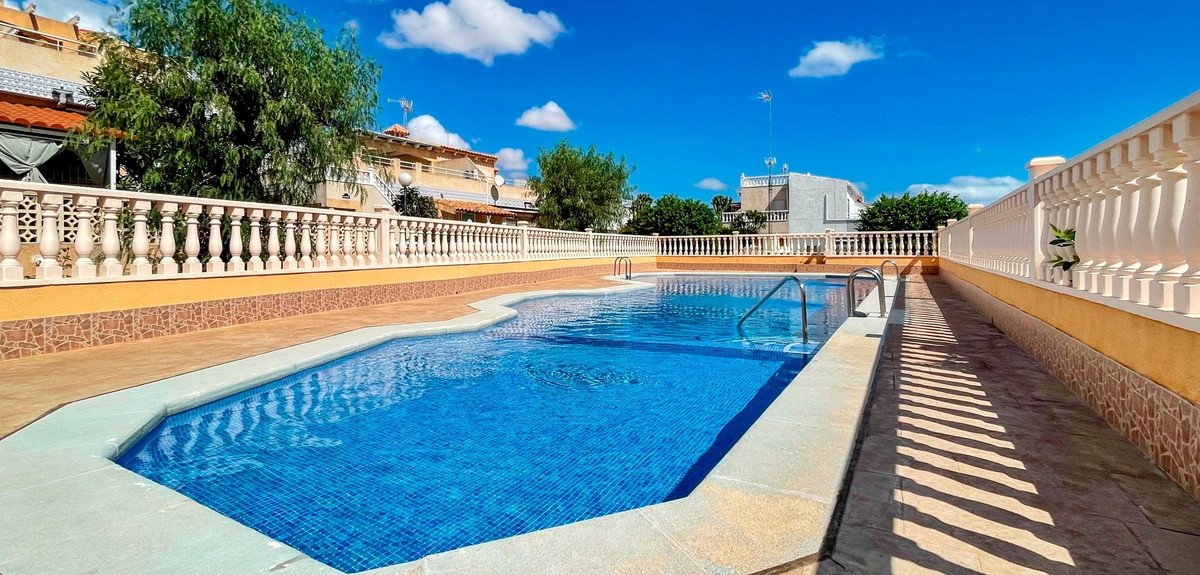 A rectangular swimming pool with blue tiled water under a bright blue sky with white clouds. Buildings in the background.