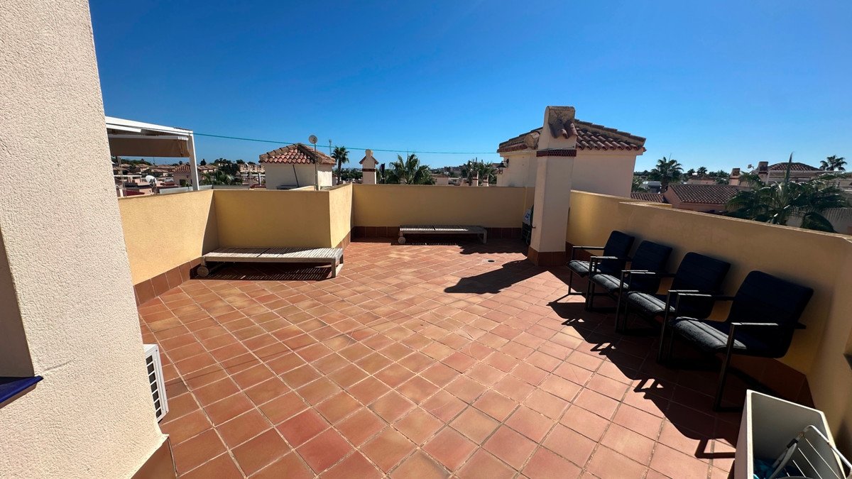A spacious tiled terrace with built-in seating and chairs, offering a view of rooftops and palm trees under a clear blue sky.