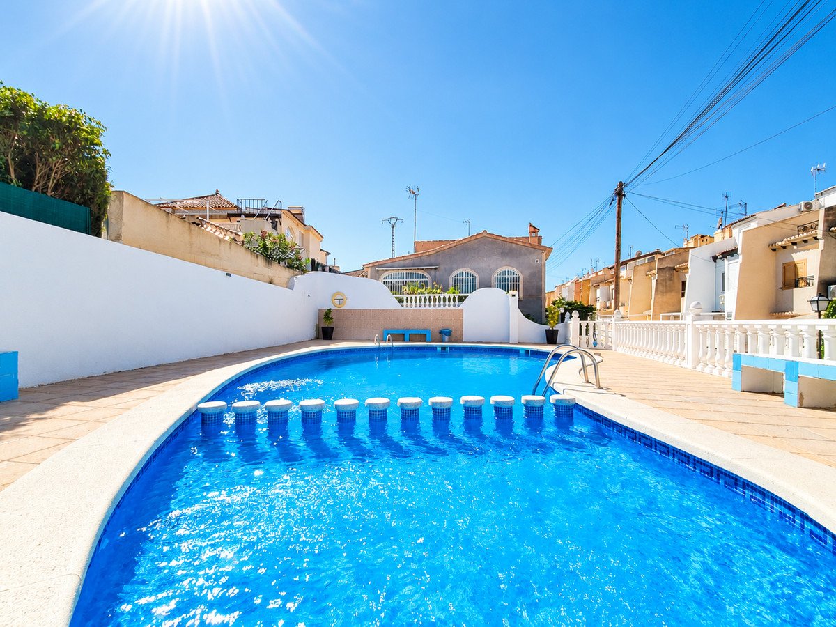 A bright blue swimming pool with a white tiled edge, surrounded by a patio and residential buildings under a clear blue sky.