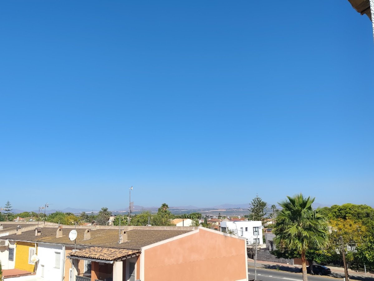 Rooftop view of houses, palm trees, and distant hills under a clear blue sky on a sunny day.