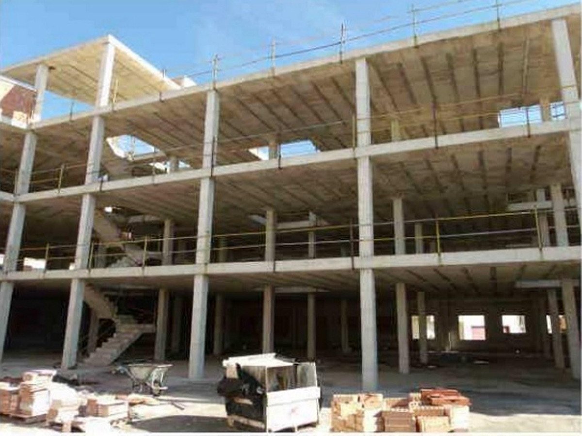 Concrete building structure under construction, showing multiple floors, support columns, and scaffolding. Materials like bricks are visible on the ground.