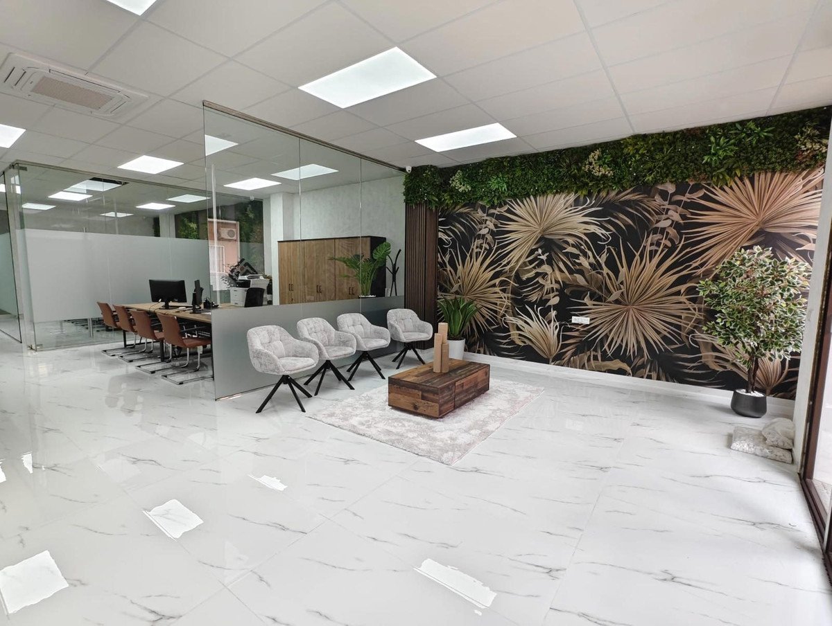 Modern office lounge area with plush chairs, a tropical leaf wallpaper accent wall, and a view into a glass-walled meeting room.