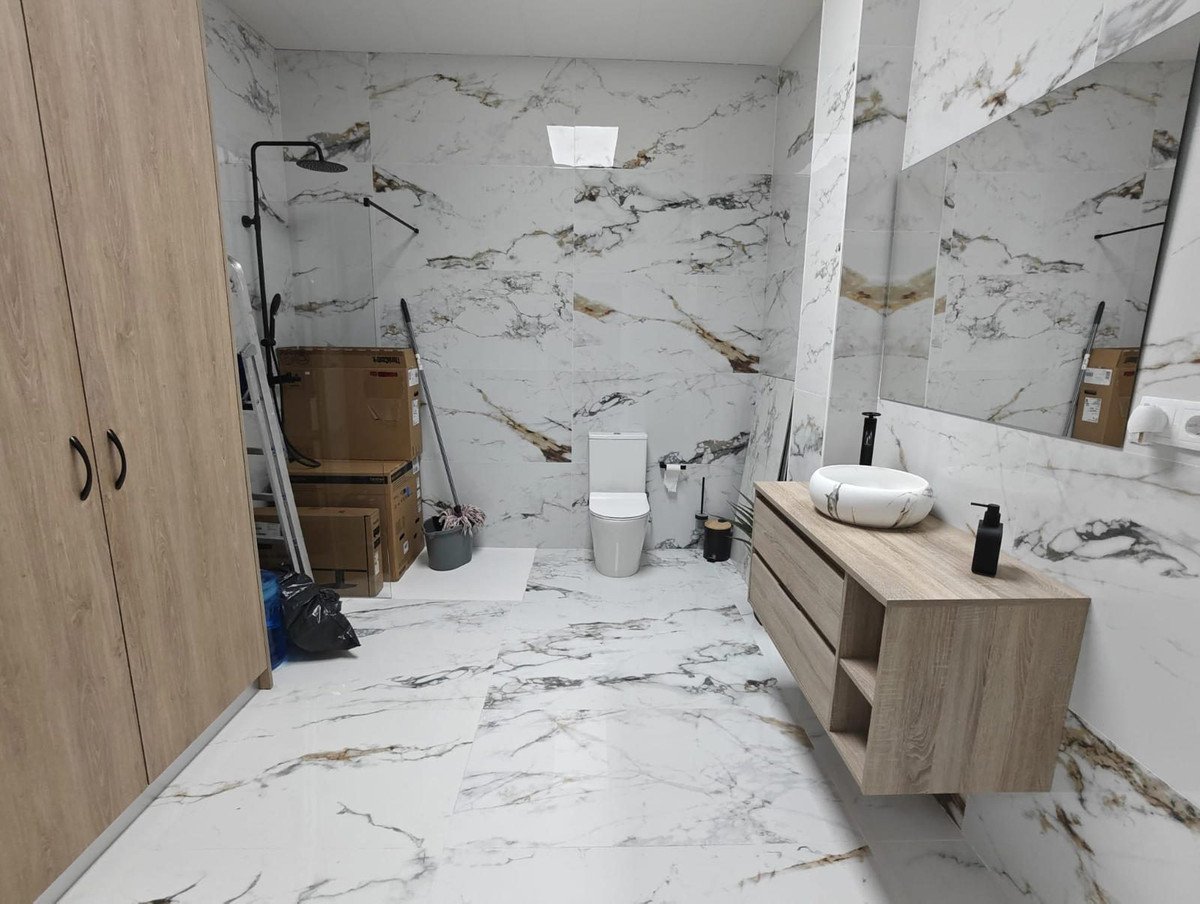 A modern bathroom with a marble-patterned vanity, sink, toilet, and a large mirror. Features a wooden cabinet and showerhead shower.